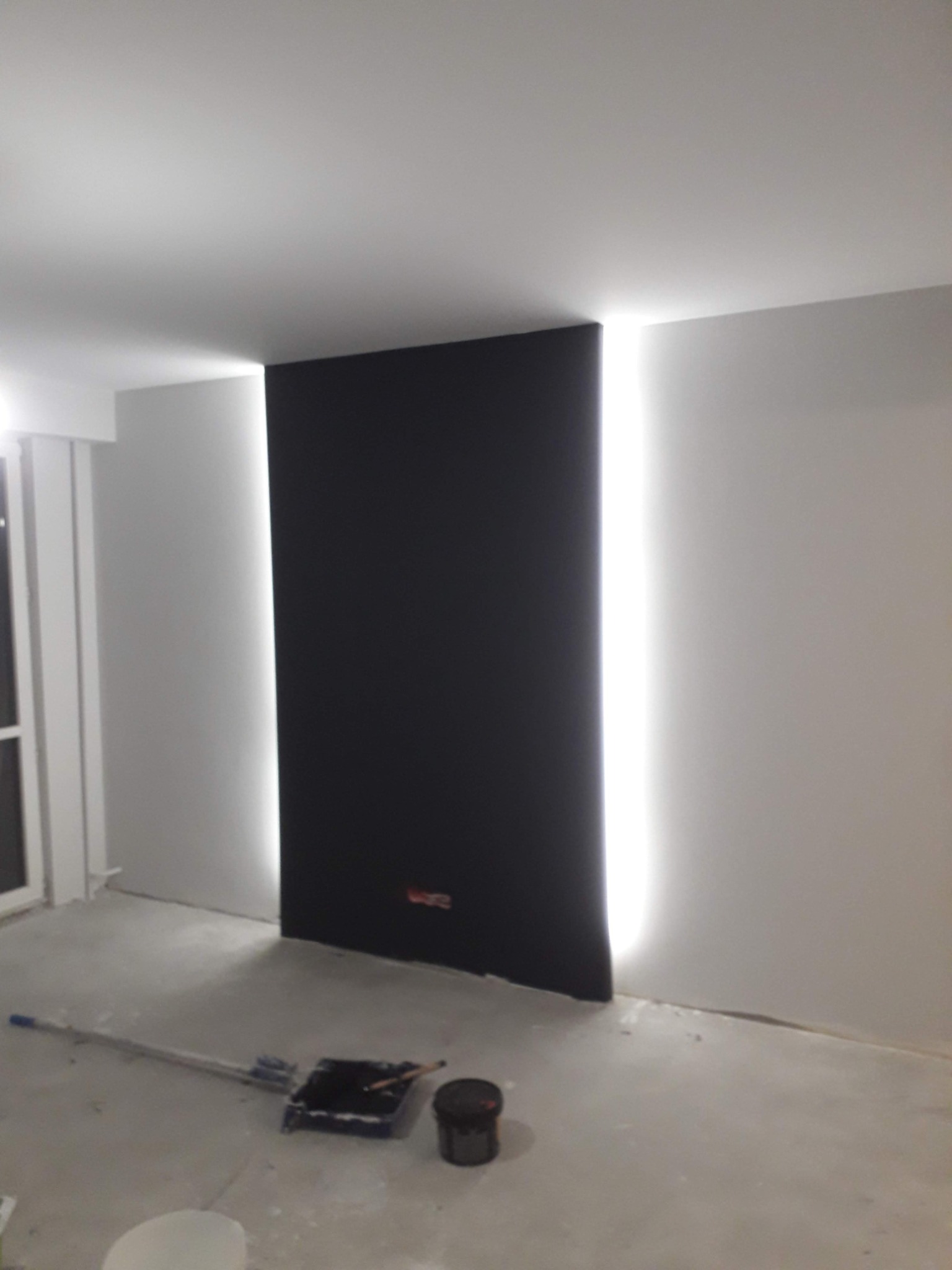 Ciemna ściana z podświetleniem LED w trakcie prac wykończeniowych, widoczny wałek i kuweta malarska na posadzce.