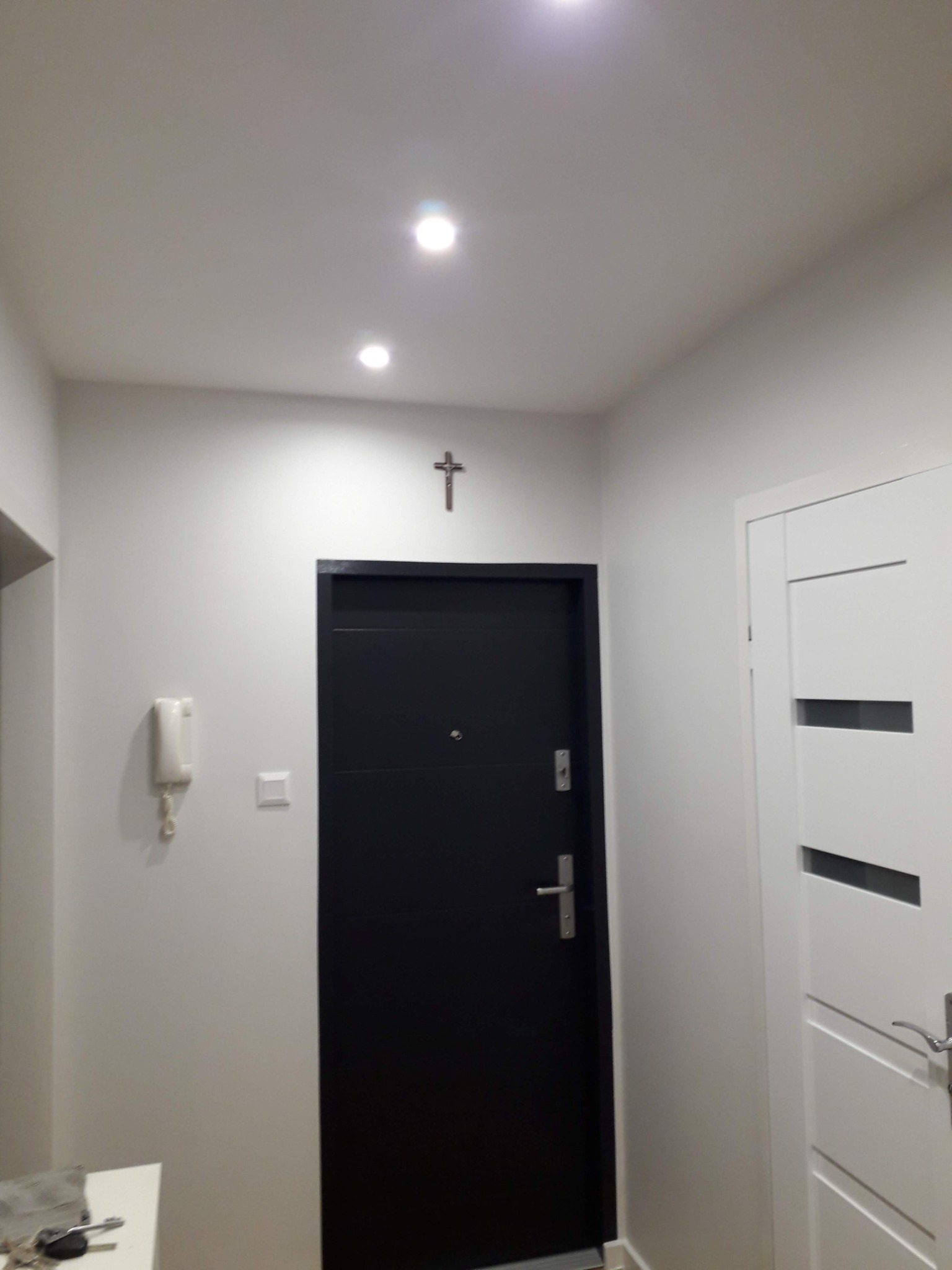 Korytarz po odświeżeniu ścian, z dwiema parami drzwi: czarne drzwi wejściowe pod krzyżem na ścianie i białe drzwi wewnętrzne z poziomymi wstawkami, oświetlenie punktowe sufitowe.