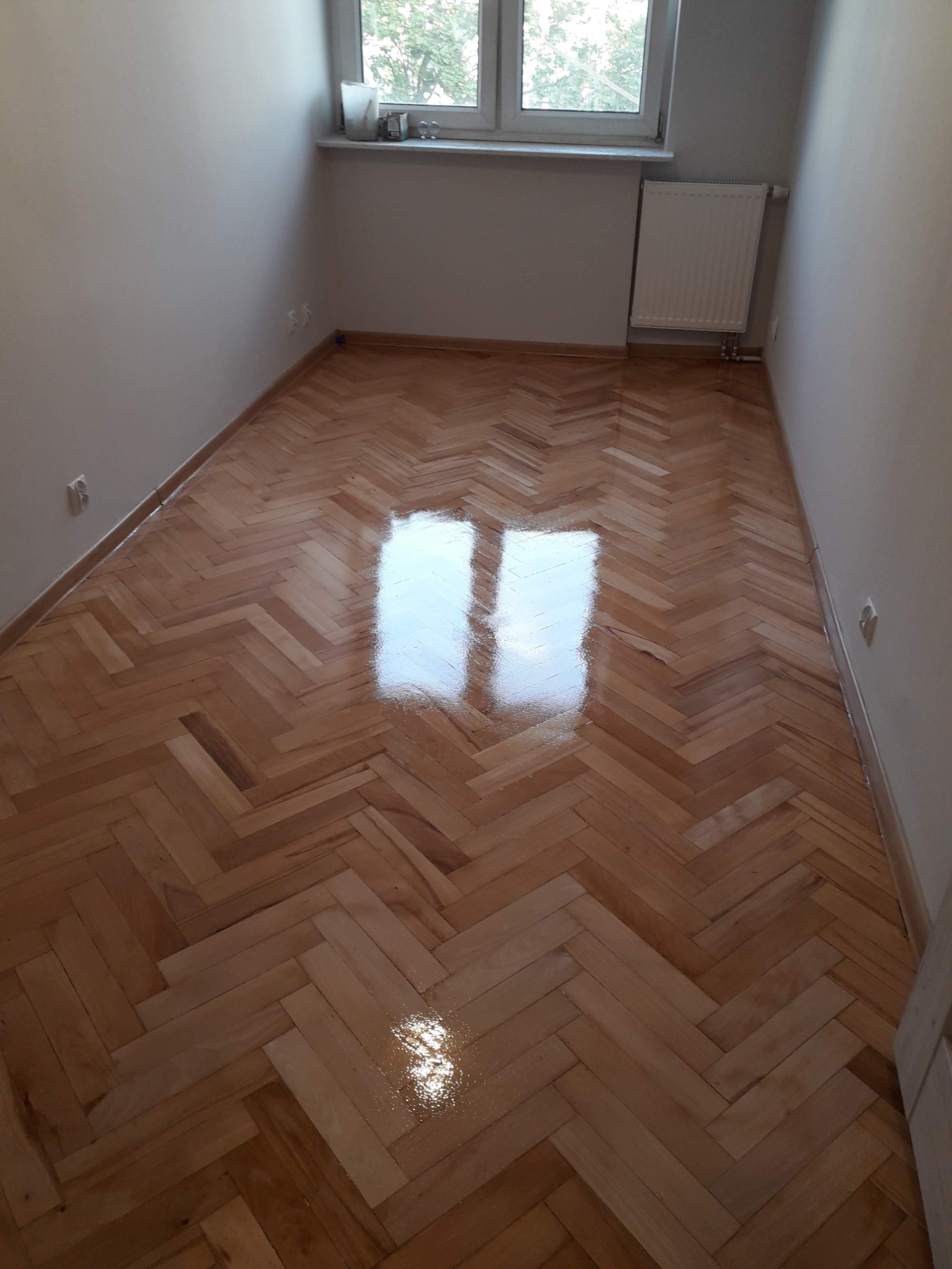 Wypolerowana, drewniana podłoga ułożona w jodełkę w pustym pokoju, z widocznym odbiciem światła z okna.