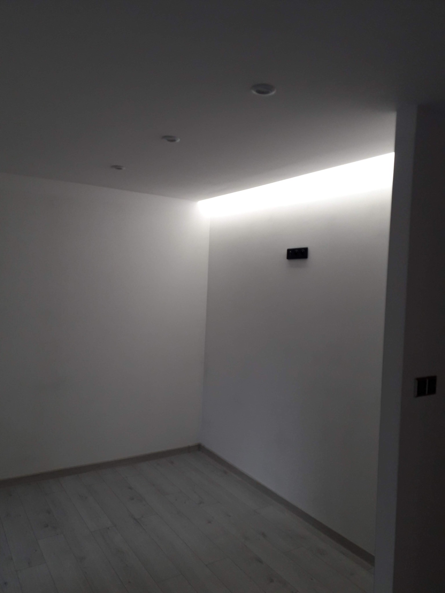 Puste, pomalowane na biało pomieszczenie z drewnianą podłogą, oświetleniem LED w suficie i gniazdkami elektrycznymi na ścianie, w trakcie wykończenia wnętrz.