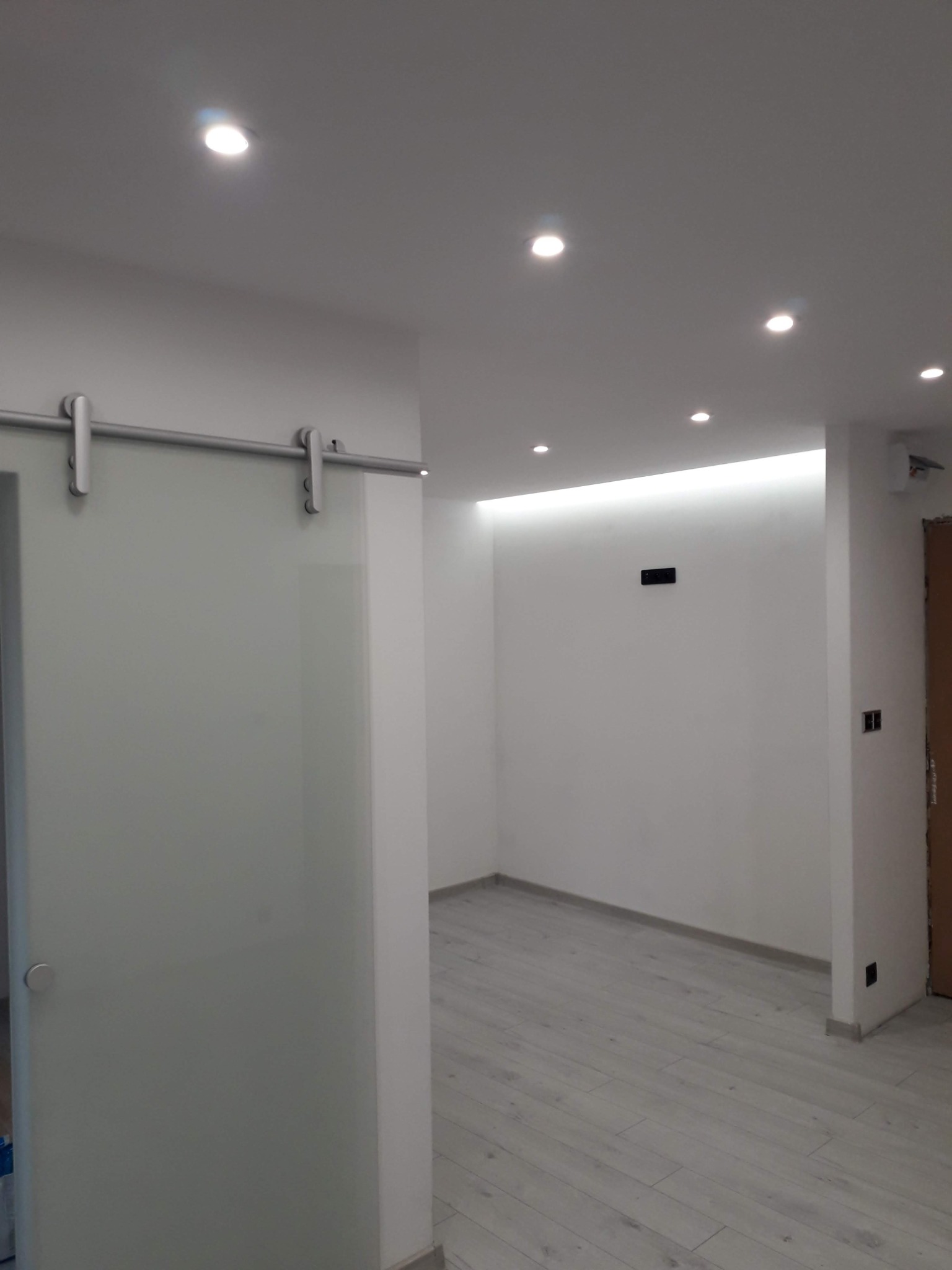 Wnętrze po remoncie: przesuwne, szklane drzwi z metalowym prowadzeniem, białe ściany z punktowym oświetleniem sufitowym i drewnopodobna podłoga.