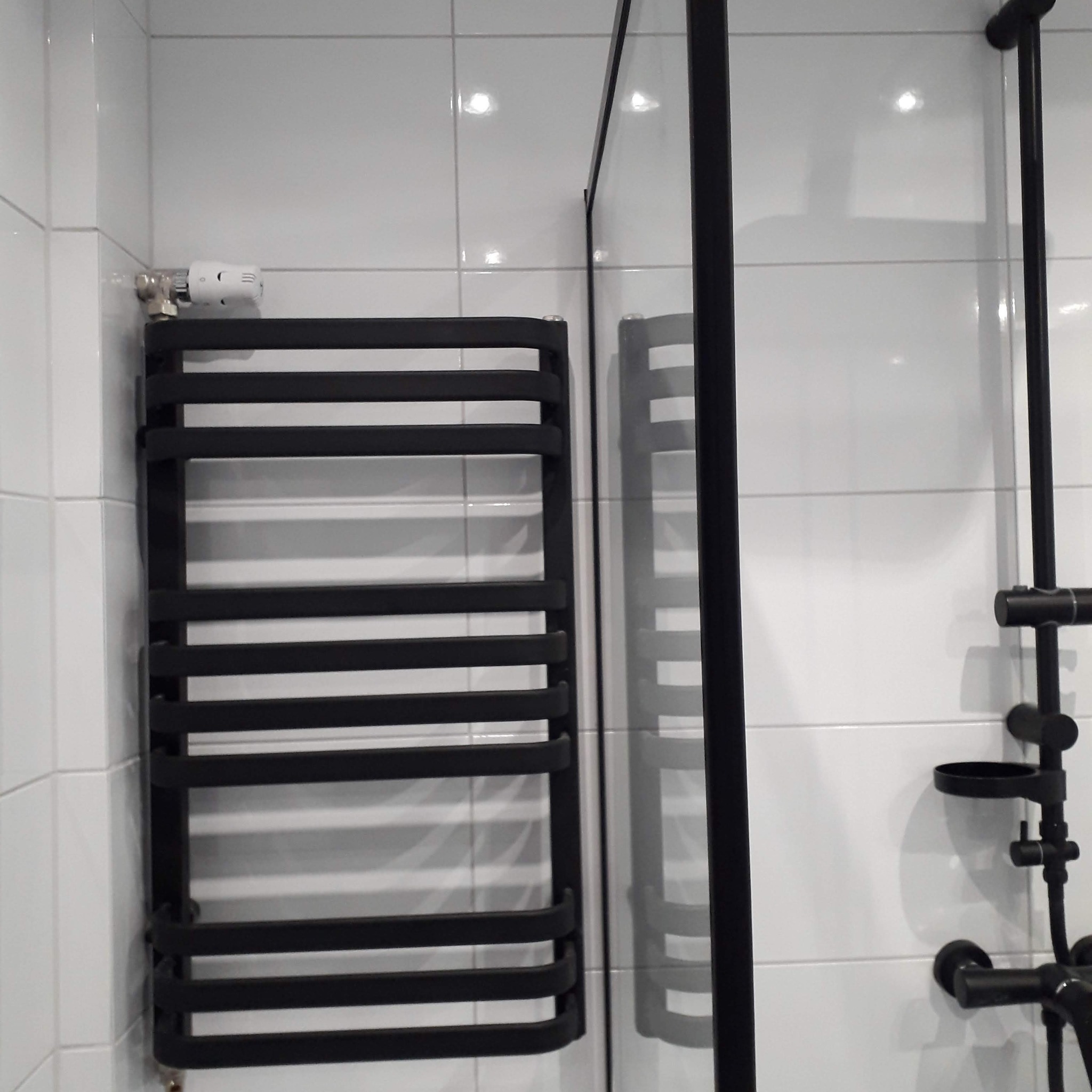 Czarny grzejnik łazienkowy drabinkowy z termostatem, obok kabina prysznicowa z czarnymi profilami i czarna bateria prysznicowa na białych, błyszczących płytkach.
