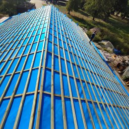 Mykhailo Mazuryk - Niebieska membrana dachowa z drewnianą konstrukcją łat, widok z góry na budowę dachu, w tle zieleń i budynki gospodarcze, dekarz przy pracy.