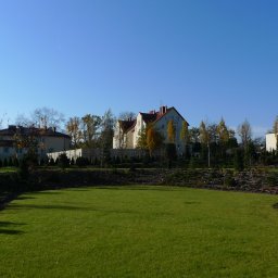 OXALIS Damian Florek - Rozległy trawnik z rolki przed domem jednorodzinnym z czerwonym dachem, otoczony ozdobnymi krzewami i drzewami, z małym oczkiem wodnym wykończonym kamieniami w prawym dolnym rogu, na tle błękitnego...