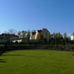 Rozległy trawnik z rolki przed domem jednorodzinnym z czerwonym dachem, otoczony ozdobnymi krzewami i drzewami, z małym oczkiem wodnym wykończonym kamieniami w prawym dolnym rogu, na tle błękitnego...