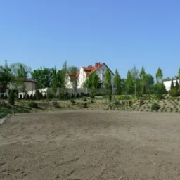 Przygotowany teren pod przyszły ogród: wyrównana ziemia z widocznymi śladami bronowania, w tle dom jednorodzinny z białymi ścianami i czerwoną dachówką, otoczony zielenią drzew i krzewów.