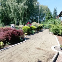 Damian Florek OXALIS - Przygotowana ścieżka ogrodowa z jasnymi obrzeżami z kostki, otoczona bujną roślinnością, w tym czerwonym klonem palmowym i różami przy żółtym domu.
