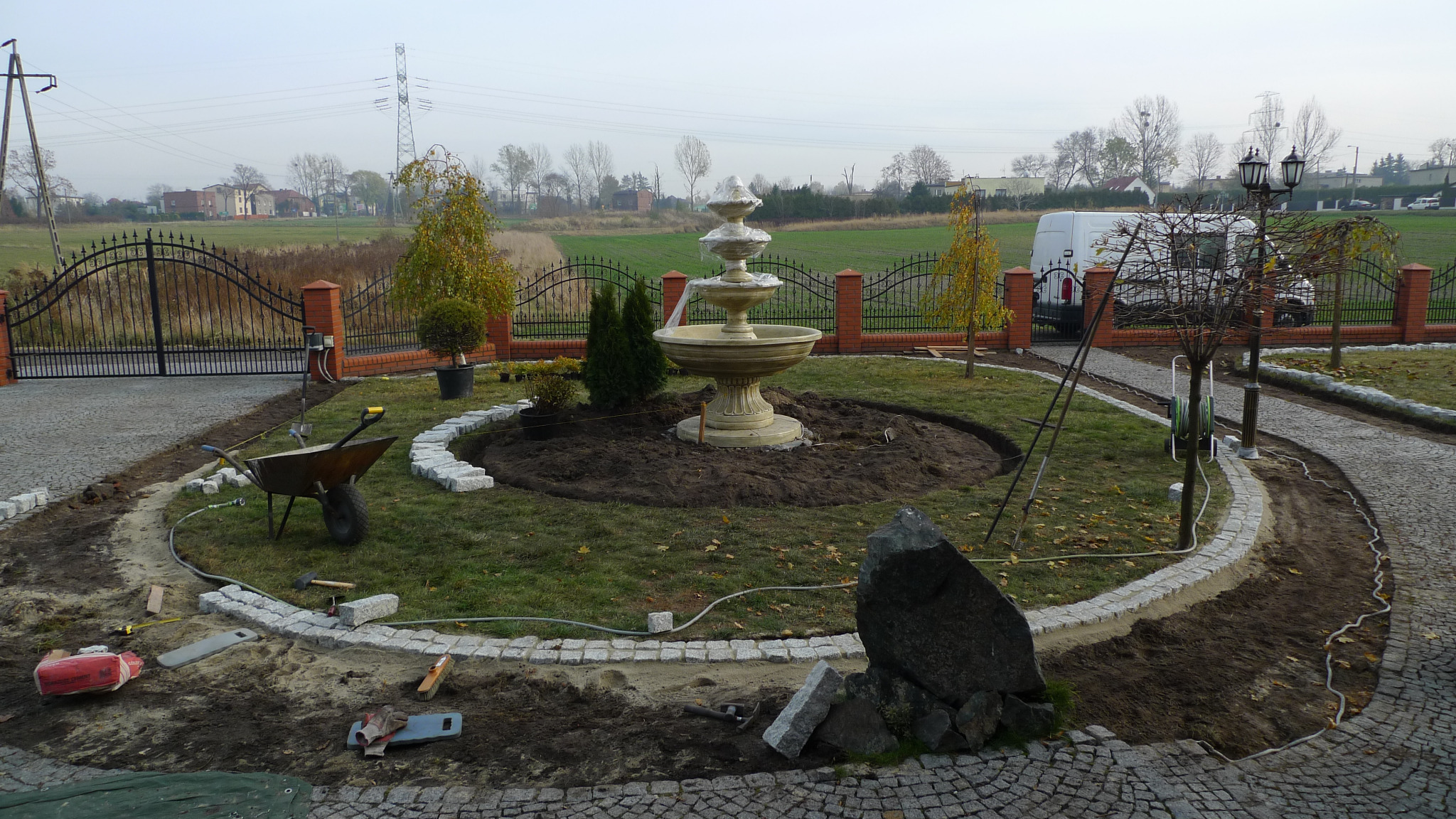 Ogród w trakcie aranżacji: fontanna otoczona ziemią i obrzeżem z kostki granitowej, widoczny wózek, narzędzia i materiały budowlane, trawa, drzewa oraz ogrodzenie z bramą w tle.