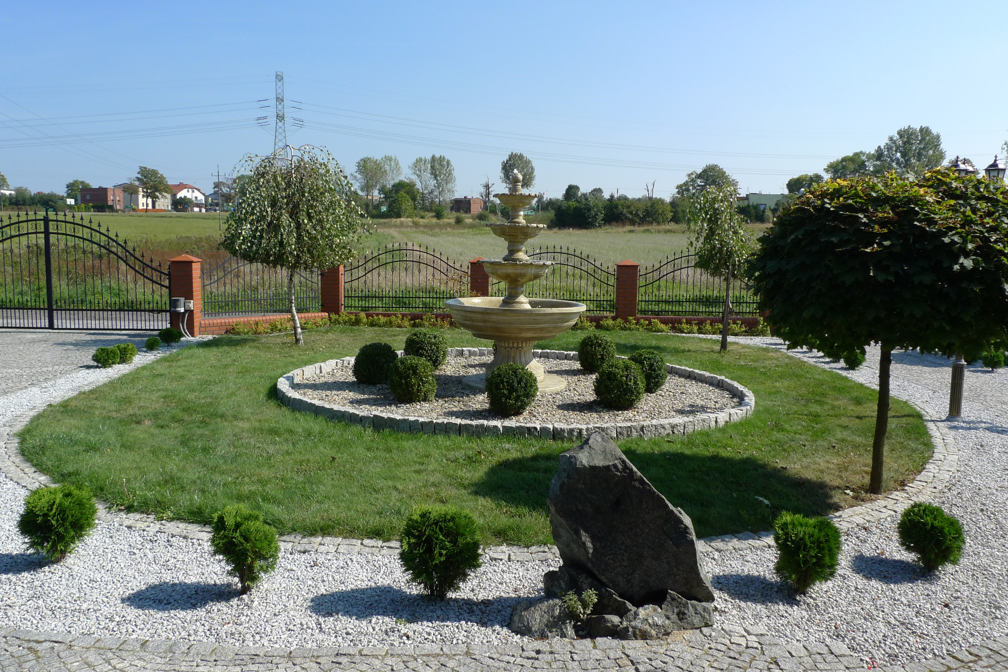 Ogród z fontanną, kulistymi krzewami i żwirową ścieżką, widok z poziomu gruntu na tle metalowego ogrodzenia i zielonego pola.