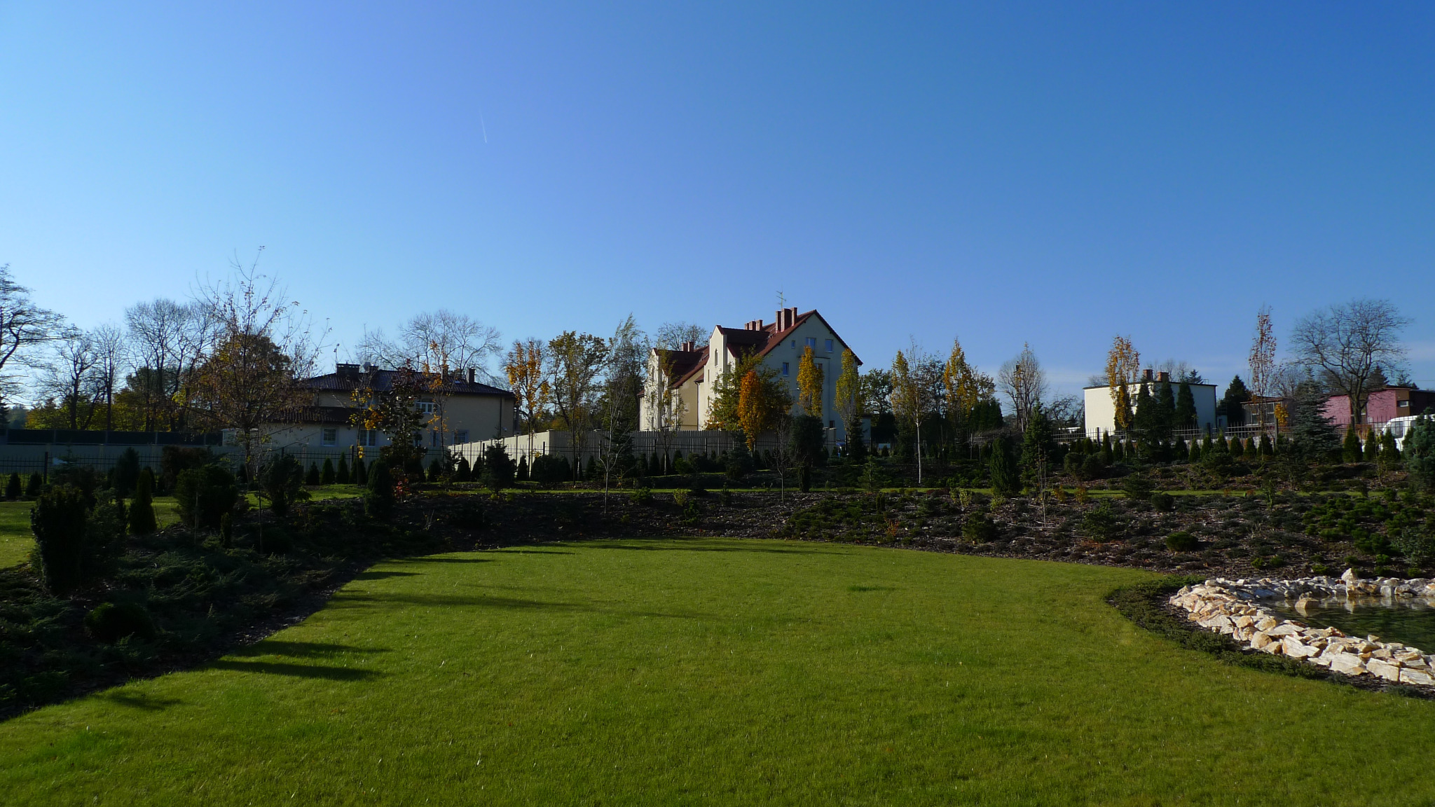 Rozległy trawnik z rolki przed domem jednorodzinnym z czerwonym dachem, otoczony ozdobnymi krzewami i drzewami, z małym oczkiem wodnym wykończonym kamieniami w prawym dolnym rogu, na tle błękitnego...