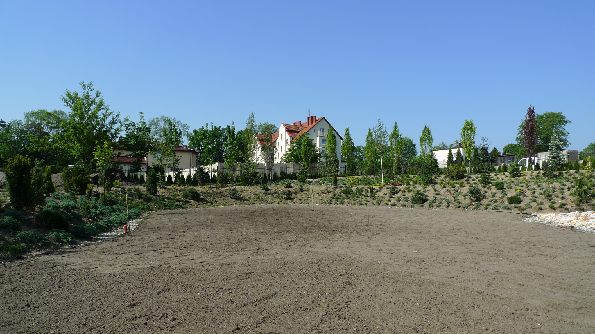 Przygotowany teren pod przyszły ogród: wyrównana ziemia z widocznymi śladami bronowania, w tle dom jednorodzinny z białymi ścianami i czerwoną dachówką, otoczony zielenią drzew i krzewów.