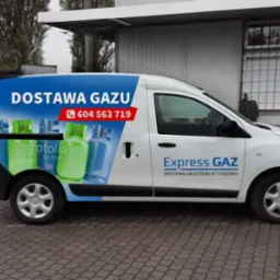 Biała furgonetka z niebieskim nadrukiem reklamowym 'Dostawa Gazu' i numerem telefonu, zaparkowana na brukowanej nawierzchni