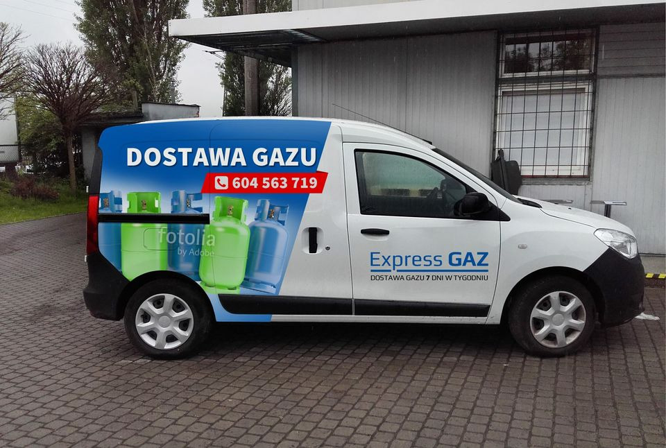 Biała furgonetka z niebieskim nadrukiem reklamowym 'Dostawa Gazu' i numerem telefonu, zaparkowana na brukowanej nawierzchni