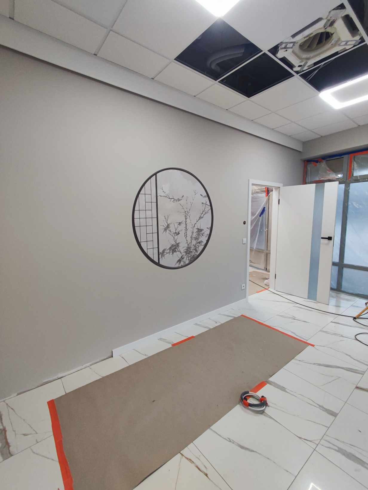 Szara ściana z okrągłą dekoracją ścienną w stylu orientalnym, zabezpieczona podłoga i listwy przypodłogowe podczas prac wykończeniowych, otwarte drzwi z niebieskim paskiem, sufit z widocznymi...