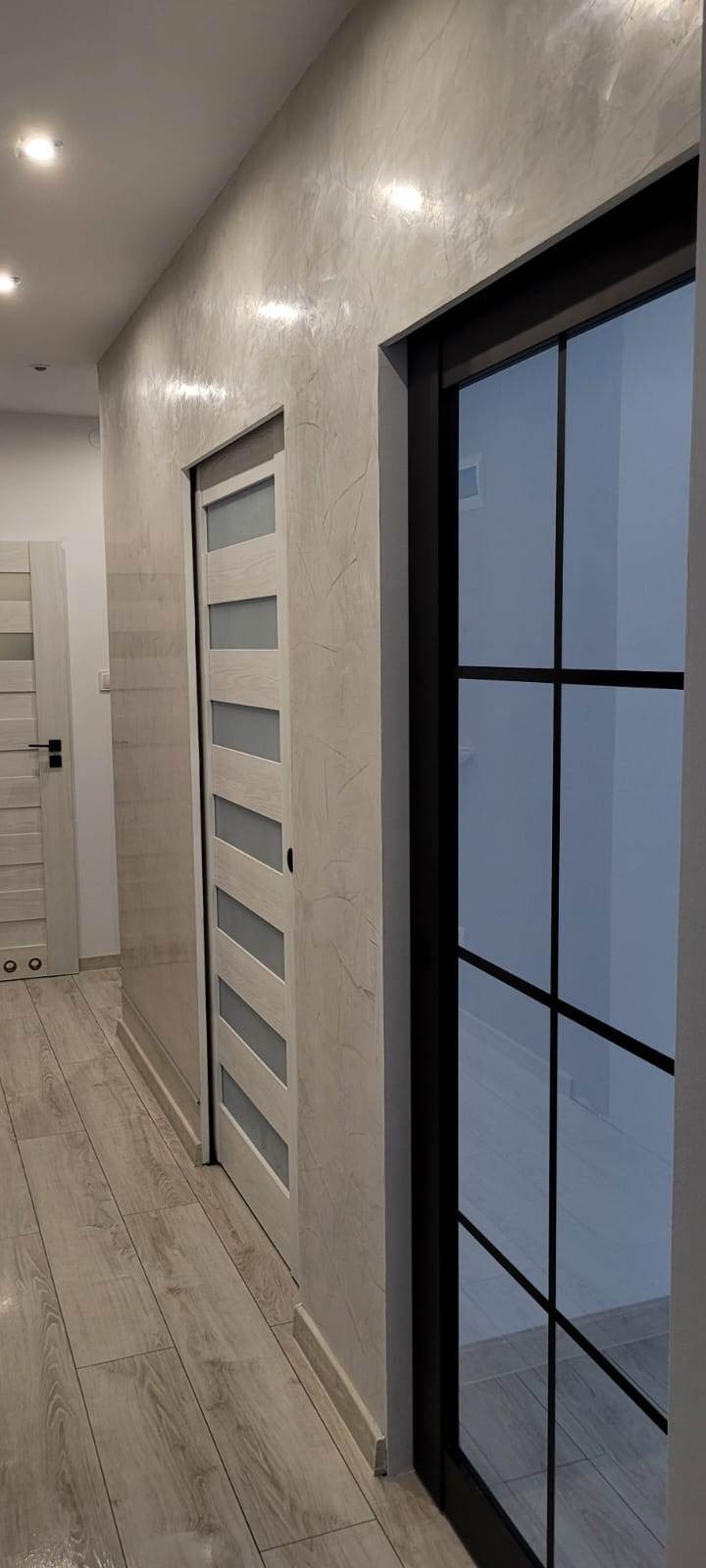 Korytarz po odświeżeniu z dwiema parami drzwi, jedna para przesuwna z poziomymi szybami, druga z czarnymi szprosami, ściany pokryte dekoracyjnym tynkiem w odcieniach szarości, podłoga wyłożona...