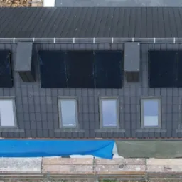 Widok z góry na dom z zainstalowanymi panelami słonecznymi na dachu pokrytym ciemną dachówką, widoczne okna dachowe, rusztowanie i częściowo osłonięty teren budowy.