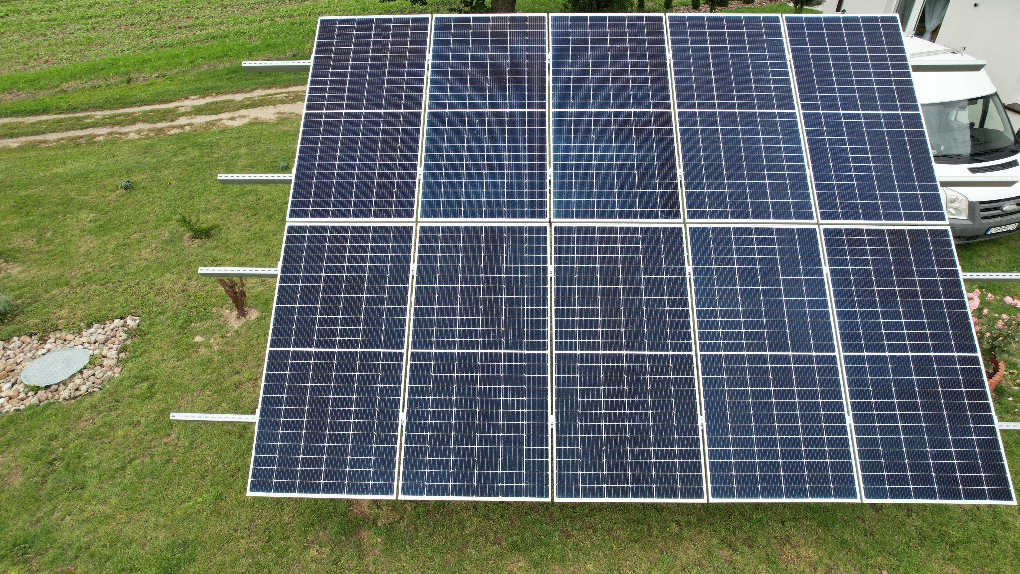 Widok z góry na zainstalowane panele słoneczne na trawniku obok białego samochodu dostawczego, widoczne elementy montażowe i dekoracyjny element z kamieni.