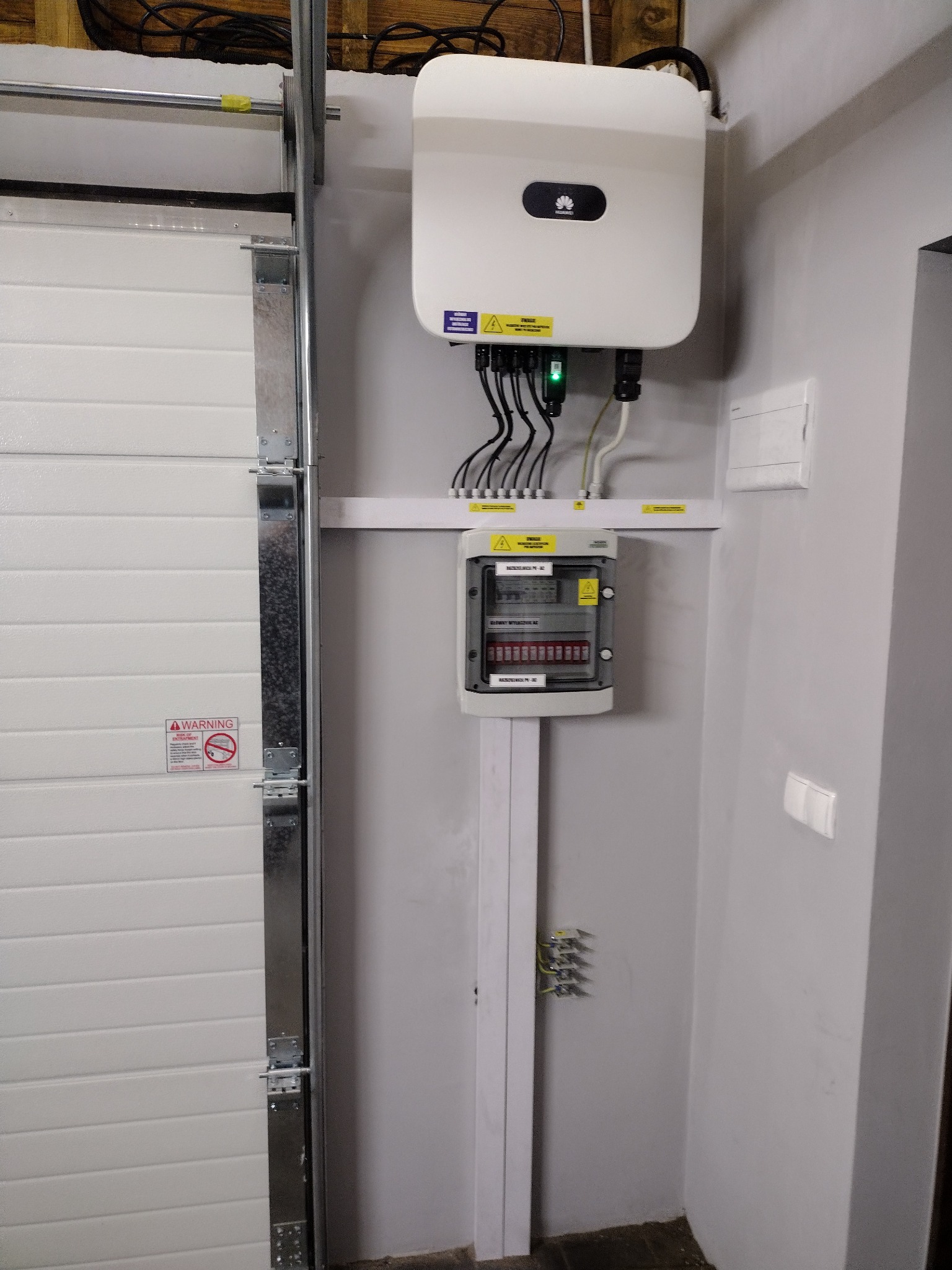 Biały inwerter Huawei zamontowany na szarej ścianie w pomieszczeniu gospodarczym, obok bramy garażowej i skrzynki z bezpiecznikami.