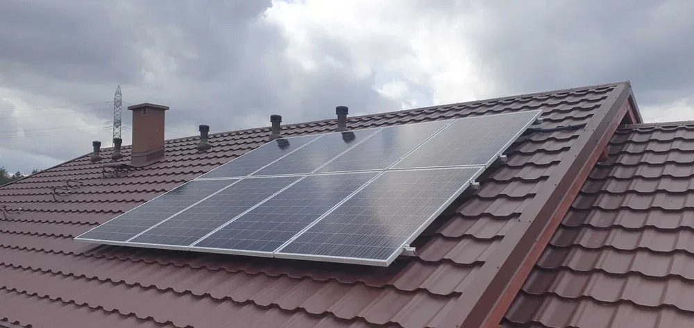Panel fotowoltaiczny zamontowany na brązowym dachu z blachodachówki, widoczny komin i odpowietrzniki dachowe na tle pochmurnego nieba.