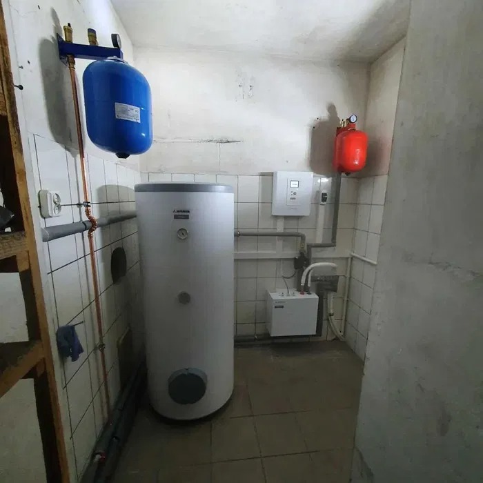 Bojler akumulacyjny z niebieskim i czerwonym zbiornikiem wyrównawczym w kotłowni wyłożonej białymi płytkami, widoczne rury miedziane i biała skrzynka sterownicza.