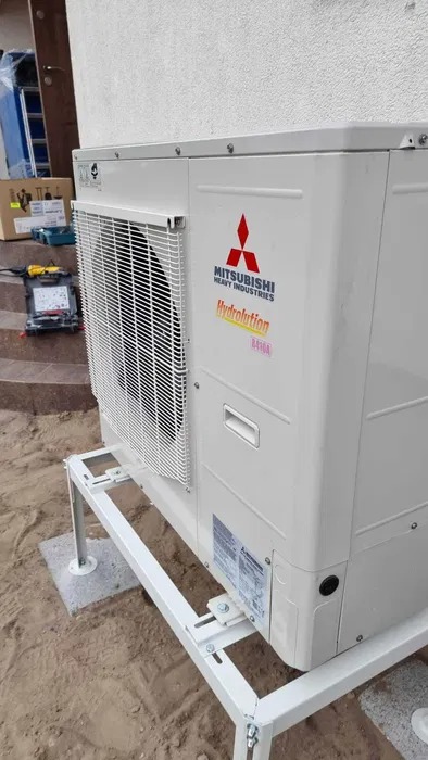 Zewnętrzna jednostka klimatyzacji Mitsubishi Heavy Industries na białym stelażu, widoczne narzędzia i karton na schodach w tle.