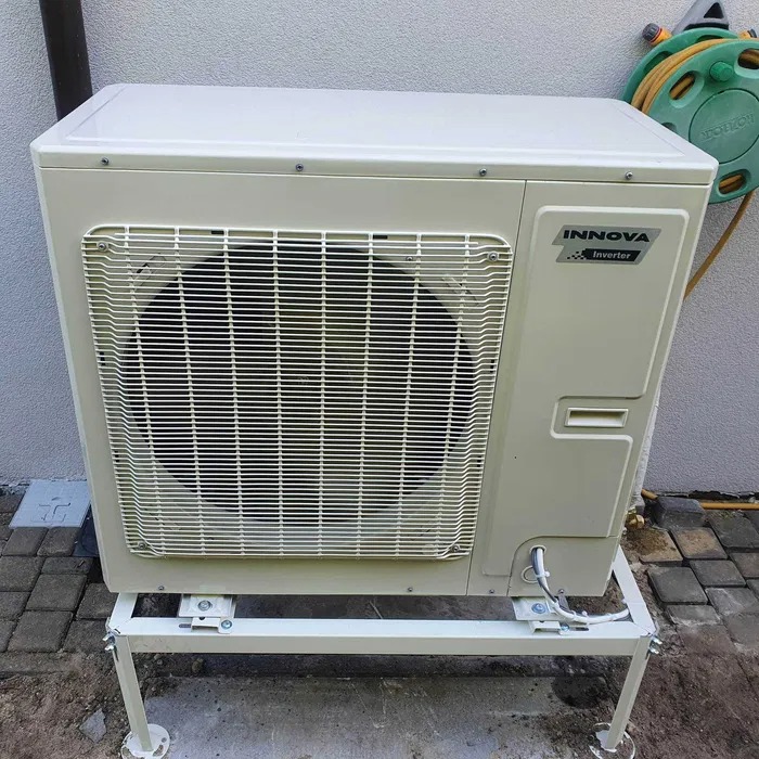 Zewnętrzna jednostka klimatyzacji Innova Inverter zamontowana na metalowym stelażu z widocznymi przewodami, na tle ściany i zwiniętego węża ogrodowego.