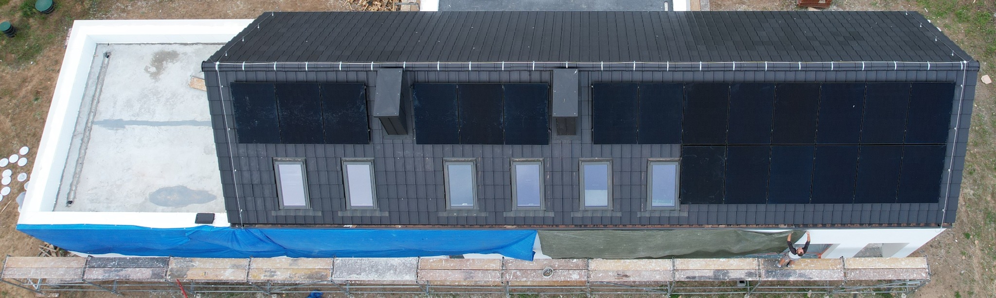 Widok z góry na dom z zainstalowanymi panelami słonecznymi na dachu pokrytym ciemną dachówką, widoczne okna dachowe, rusztowanie i częściowo osłonięty teren budowy.