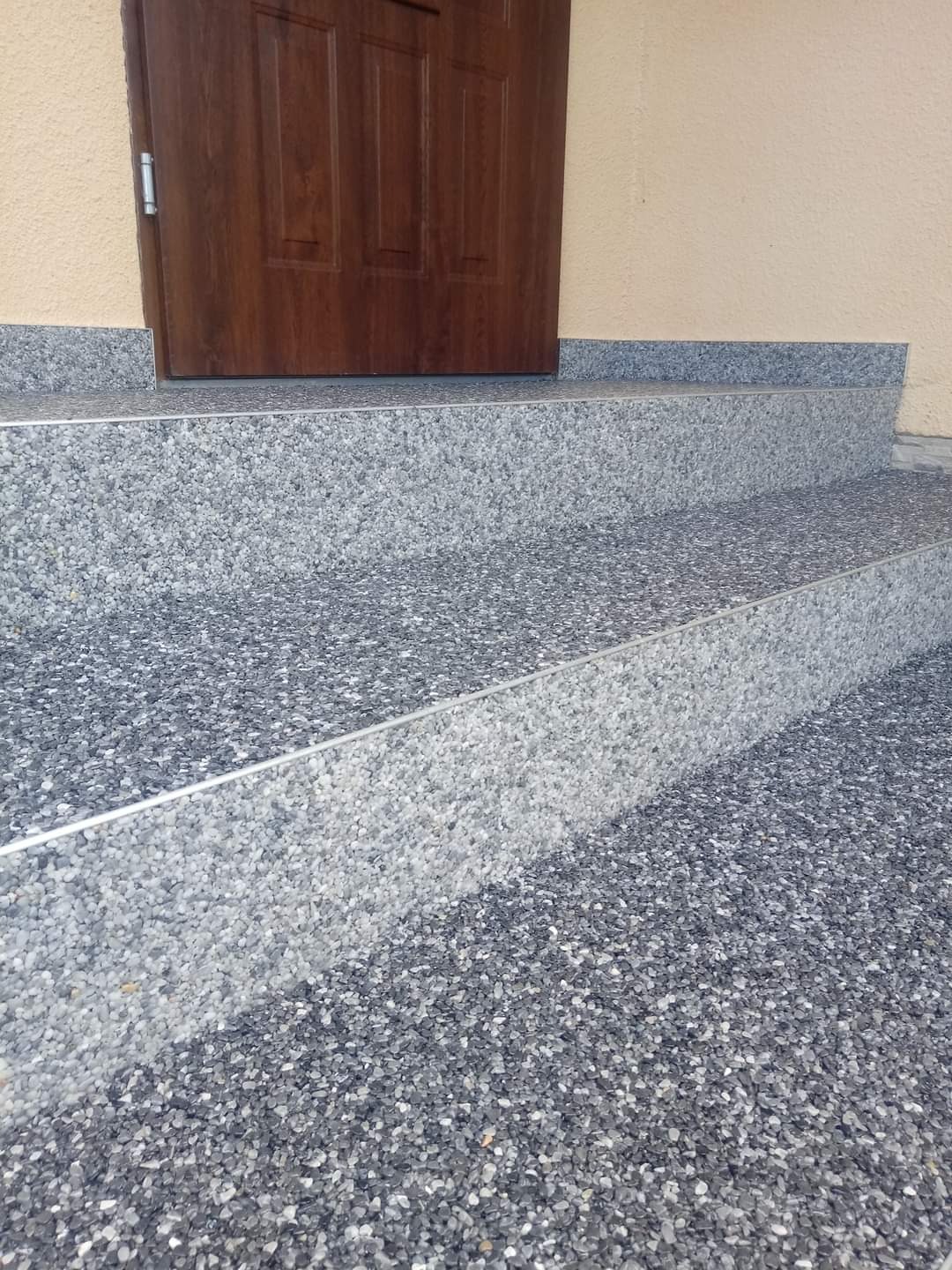 Schody wejściowe pokryte szarą, kamyczkową posadzką żywiczną z metalowymi listwami wykończeniowymi, prowadzące do drewnianych drzwi wejściowych.