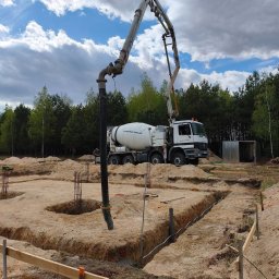 Ever-Bud Stasiak - Fundamenty domu w trakcie budowy: betonowanie z użyciem pompy do betonu, widoczne zbrojenia i szalunki. W tle las i błękitne niebo z chmurami.