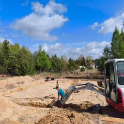 Ever-Bud Stasiak - Wykonawca w trakcie prac ziemnych pod fundamenty domu jednorodzinnego, używając minikoparki Takeuchi TB216. Widoczne wykopane rowy i piasek. Błękitne niebo z chmurami w tle.