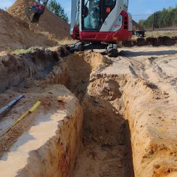Ever-Bud Stasiak - Mini koparka Takeuchi TB216 w trakcie prac ziemnych, kopania rowu pod fundamenty. Widoczne ślady opon i wykopany rów w piaszczystej glebie. Poziomica na krawędzi wykopu.