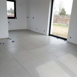 Ever-Bud Stasiak - Jasnoszara, duża płytka podłogowa w minimalistycznym wnętrzu z dużym oknem i małymi, niebieskimi krzyżykami dystansowymi. Widok na zewnątrz.