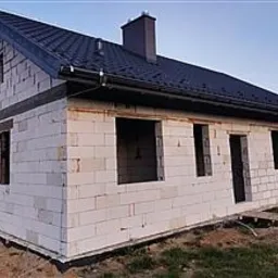 Surowy stan domu jednorodzinnego z białego bloczka, z czarnym dachem i kominem, widoczne otwory okienne i drzwiowe, teren wokół niezagospodarowany.