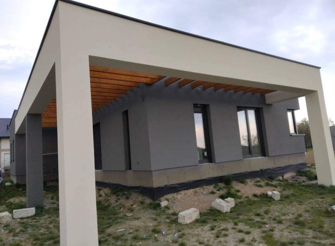 Nowoczesny dom jednorodzinny z szarą elewacją i jasnymi elementami konstrukcyjnymi, widoczne drewniane belki pod dachem, teren wokół domu w trakcie zagospodarowywania.