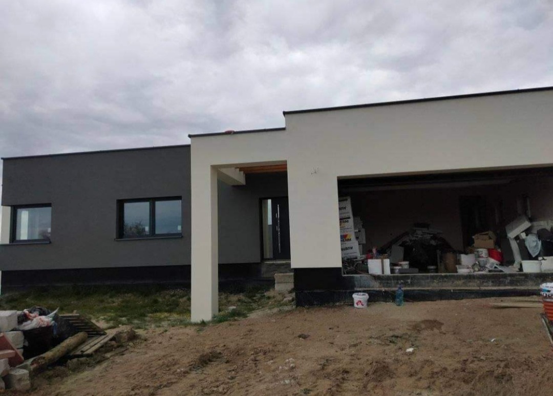 Nowoczesny, parterowy dom z płaskim dachem w trakcie wykończenia elewacji, z widocznymi materiałami budowlanymi w otwartym garażu, część budynku w kolorze szarym, część w jasnym beżu, na pierwszym...