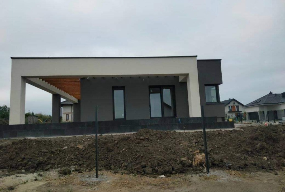 Nowoczesny dom jednorodzinny z płaskim dachem, szarą elewacją i drewnianą pergolą nad tarasem, widoczny w trakcie prac wykończeniowych.