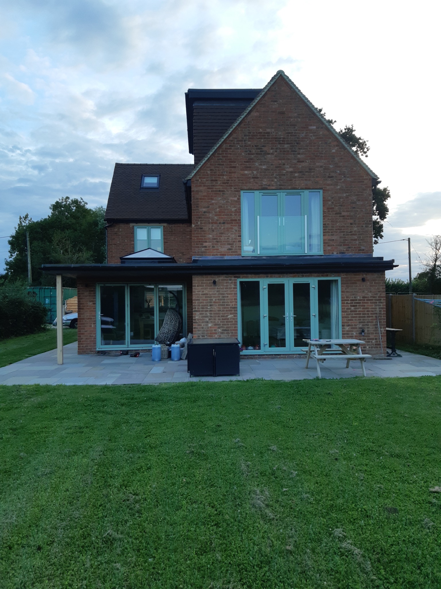 Dwukondygnacyjny dom z czerwonej cegły z dobudowaną częścią, widok z ogrodu, z zielonymi ramami okiennymi i drzwiowymi, oraz szarym tarasem.