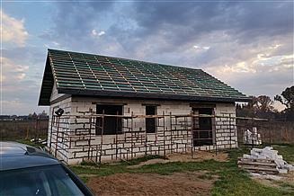 Surowy stan domu z białego pustaka, z zielonym dachem z dachówki ceramicznej, widoczne rusztowania i paleta z bloczkami, pochmurne niebo w tle.