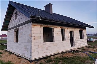 Surowy stan domu jednorodzinnego z białego bloczka, z czarnym dachem i kominem, widoczne otwory okienne i drzwiowe, teren wokół niezagospodarowany.