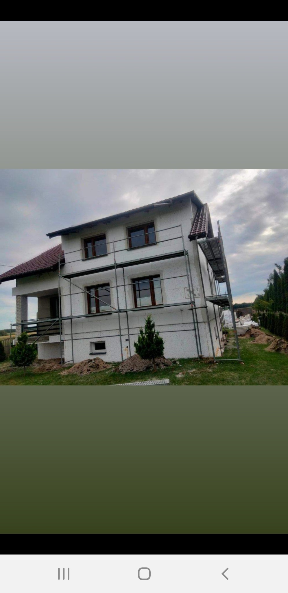 Dom murowany w trakcie ocieplania elewacji, z widocznymi rusztowaniami i częściowo ocieplonymi ścianami, na tle pochmurnego nieba.