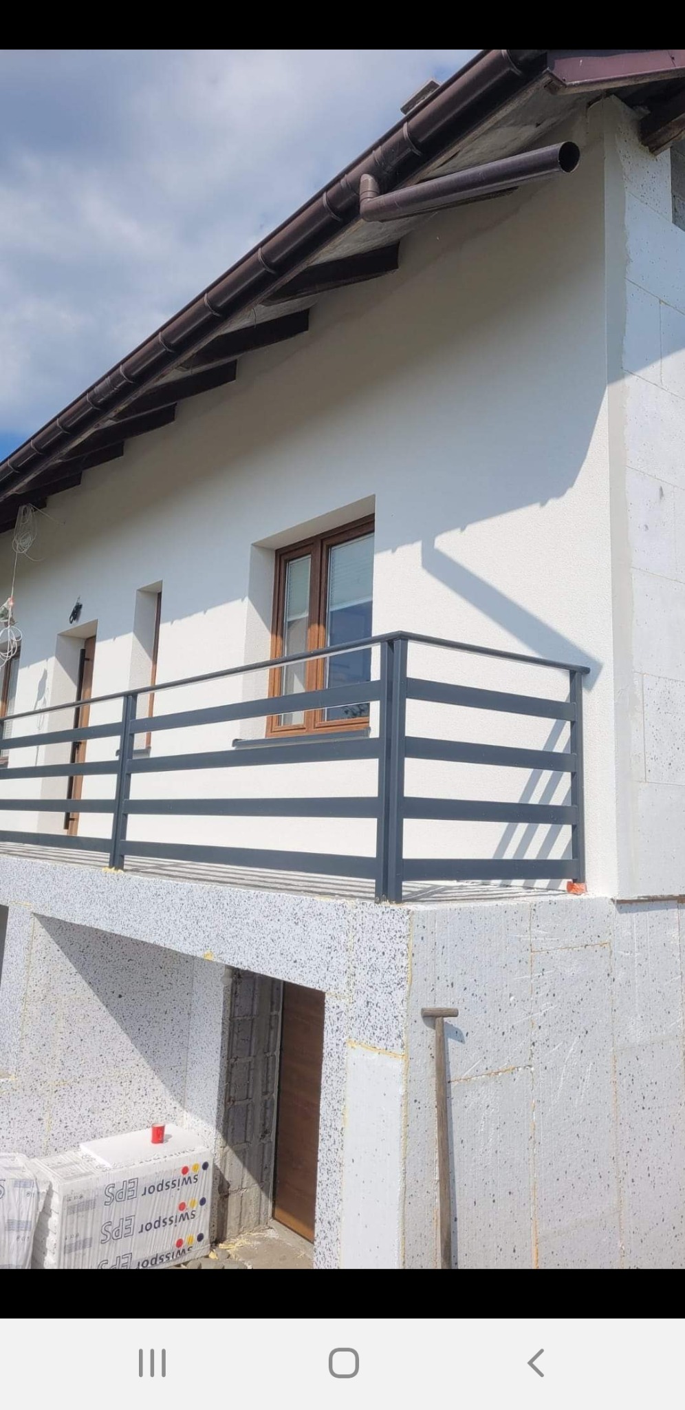 Niedokończony murowany budynek z ociepleniem elewacji, widoczne pakiety styropianu EPS, szara balustrada balkonowa i brązowe rynny.