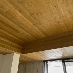 AR-DECOR WYKOŃCZENIA WNĘTRZ ARKADIUSZ PARZYGNAT - Drewniany sufit z desek w odcieniach brązu, widoczny pod kątem. Fragment ściany z oknem w tle. Widoczne niedoskonałości surowego stanu wykończenia.