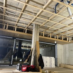 AR-DECOR WYKOŃCZENIA WNĘTRZ ARKADIUSZ PARZYGNAT - Drewniana konstrukcja sufitu podwieszanego z widocznymi instalacjami elektrycznymi i wentylacyjnymi. W tle okna i materiały budowlane. Etap prac wykończeniowych.
