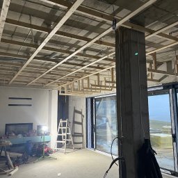 AR-DECOR WYKOŃCZENIA WNĘTRZ ARKADIUSZ PARZYGNAT - Wnętrze w trakcie prac wykończeniowych: drewniana konstrukcja sufitu podwieszanego, surowy betonowy filar, okna przesuwne. Widoczne narzędzia i drabina.