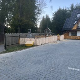AR-DECOR WYKOŃCZENIA WNĘTRZ ARKADIUSZ PARZYGNAT - Nowe, surowe drewniane ogrodzenie posesji z bramką, na tle brukowanej nawierzchni i domu z panelami słonecznymi. Widoczne drzewa i elementy placu zabaw.
