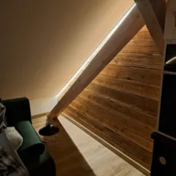 Drewniana ściana skośna na poddaszu, oświetlona listwą LED, w tle ciemnozielona sofa i lampka druciana.