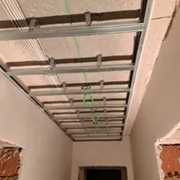 Konstrukcja metalowa podwieszanego sufitu z widocznymi przewodami elektrycznymi w zielonej izolacji, zamocowana do surowego stropu w trakcie prac wykończeniowych.