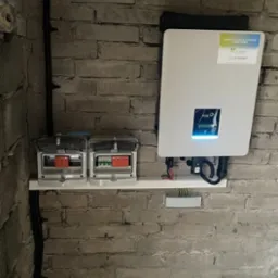 Szary mur z cegły, na nim zamontowany biały inwerter fotowoltaiczny FoxESS z wyświetlaczem i naklejką informacyjną oraz dwie skrzynki elektryczne z otwartymi klapkami, kable i przewody.