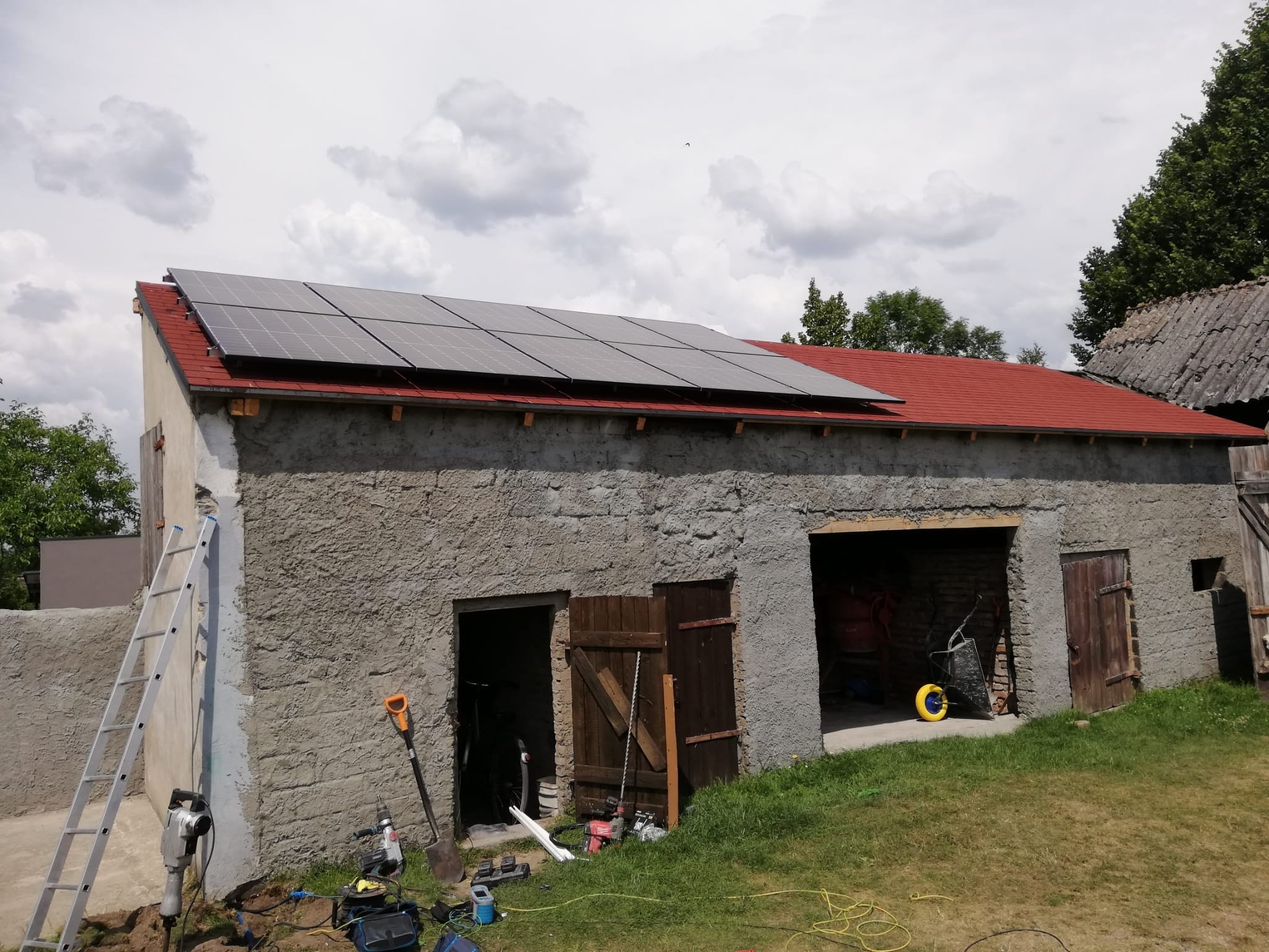 Montaż paneli fotowoltaicznych na dachu starego, kamiennego budynku gospodarczego z czerwoną dachówką, widoczna drabina i narzędzia na ziemi.