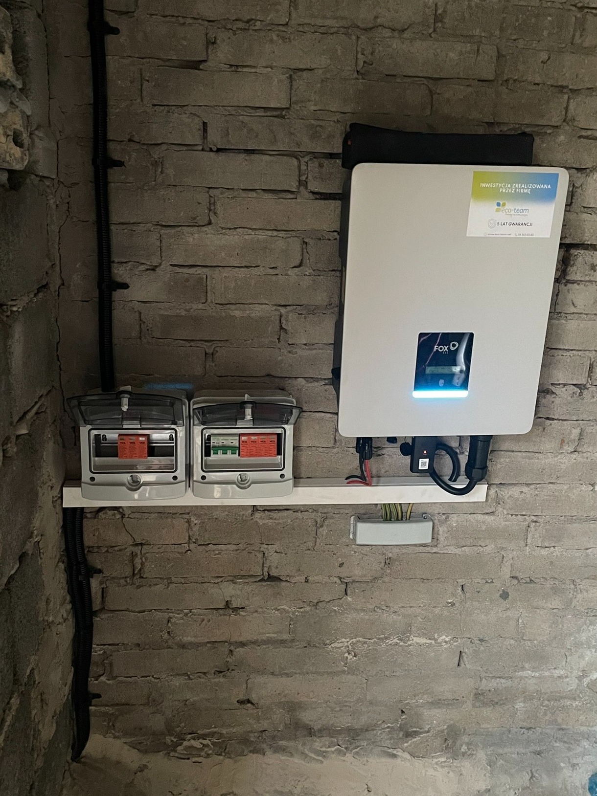Szary mur z cegły, na nim zamontowany biały inwerter fotowoltaiczny FoxESS z wyświetlaczem i naklejką informacyjną oraz dwie skrzynki elektryczne z otwartymi klapkami, kable i przewody.